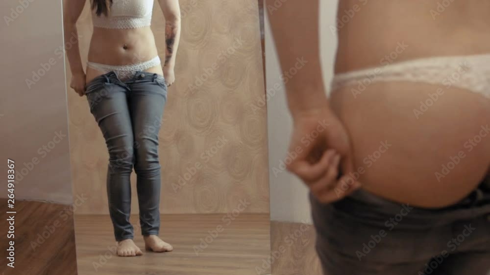 A young girl puts on jeans in front of a mirror. She can't wear jeans. The girl suffers from mild obesity. Static camera