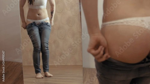A young girl puts on jeans in front of a mirror. She can't wear jeans. The girl suffers from mild obesity. Static camera