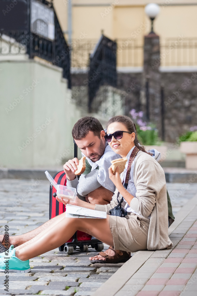 Romantic Caucasian Couple Traveling Around City. Sitting on Floor With Luggage, Camera and Map and Having Lunch with Sandwiches.