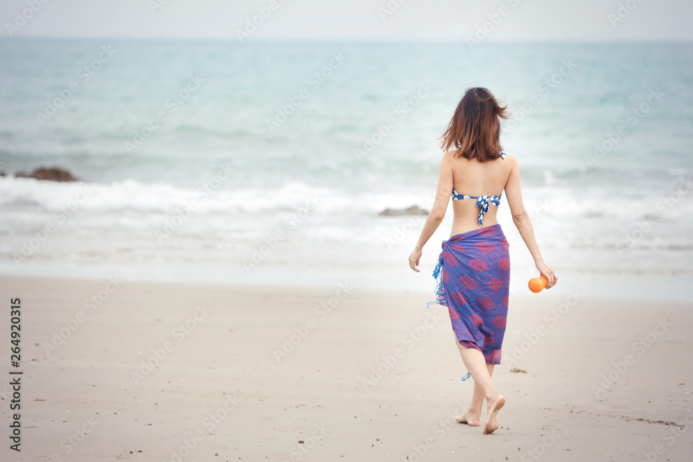 Back view of shapely beautiful girl walking on tropical beach in swimsuit and holding orange juice in nature with blue sky