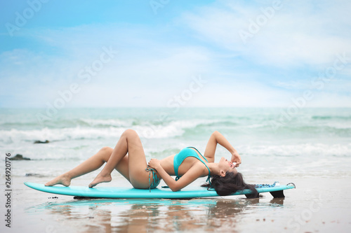 beautiful and sexy surfer girl lying on surf board on tropical sea in green swimsuit on sandy beach while she holding sunglasses during summer time with happy face. (water sport or vacation concept)