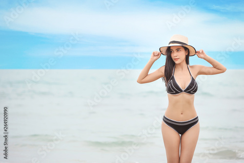 portrait of young asian beautiful and sexy woman in black bikini and white hat with beauty face standing on sandy beach with blue sky and left copy space