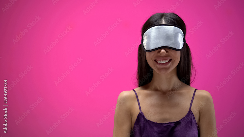 Charming female wearing eye mask for sleeping and smiling on pink background Charming female wearing eye mask for sleeping and smiling on pink background