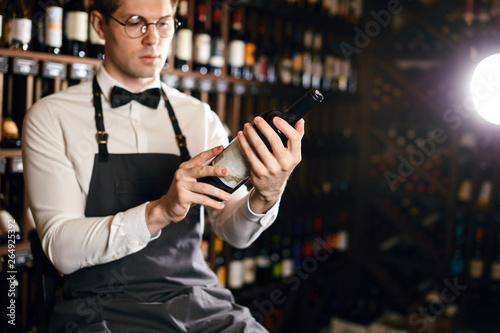 Young caucasian cavist dressed in white shirt and bowtie working in big vine shop presenting a bottle of red wine to customer photo