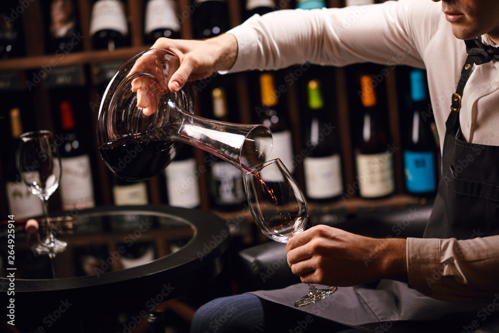 Naklejka premium Wine taster or degustator mixing red wine into carafe to make perfect color on background with wine bottles racks in wine house.
