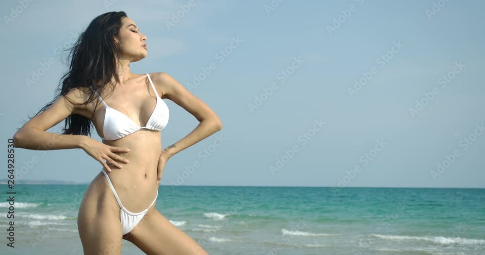 Sexy hot asian woman in white bikini posing on blue sea and sandy beach landscape. Slow motion. Summer, Travel and Beauty concept. Sexy hot asian woman in white bikini posing on blue sea and sandy beach landscape. Slow motion. Summer, Travel and Beauty concept.