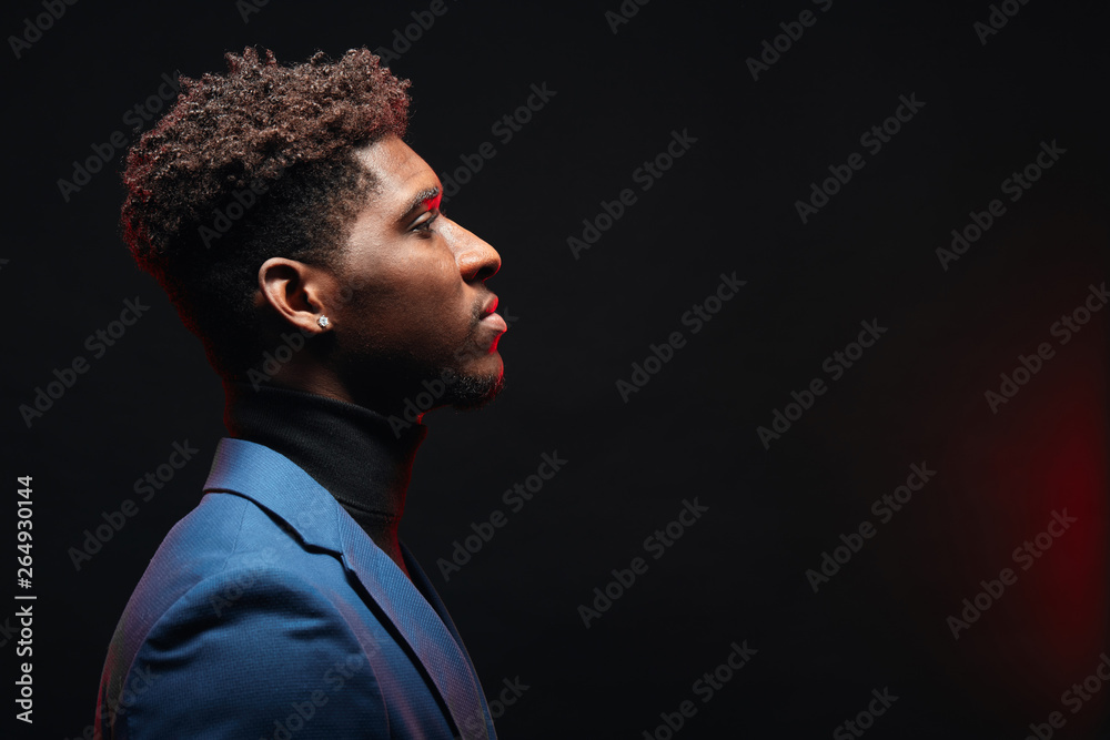 Obraz premium Side view of african handsome young man with hairstyle having neutral calm face expression standing in formal jacket on dark studio background
