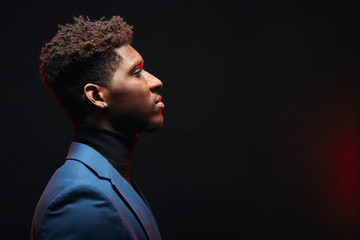 Side view of african handsome young man with hairstyle having neutral calm face expression standing in formal jacket on dark studio background