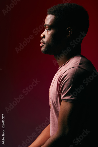 Young serene African male model with athletic body dressed in casual t-shirt posing on dark red background in profile