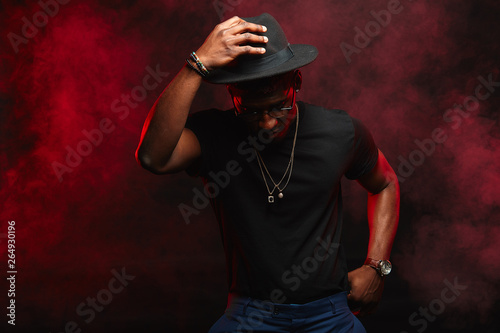 Portrait of single isolated young african hipster guy dancing in trendy hat and black t-shirt in nightclub with smoky air and darkness behind © alfa27