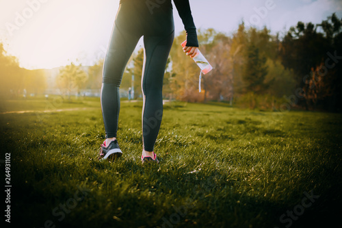 young girl feet in snickers standing in park and making running exercise on sunset and holding bottle of water in hand - fitness sport health concept