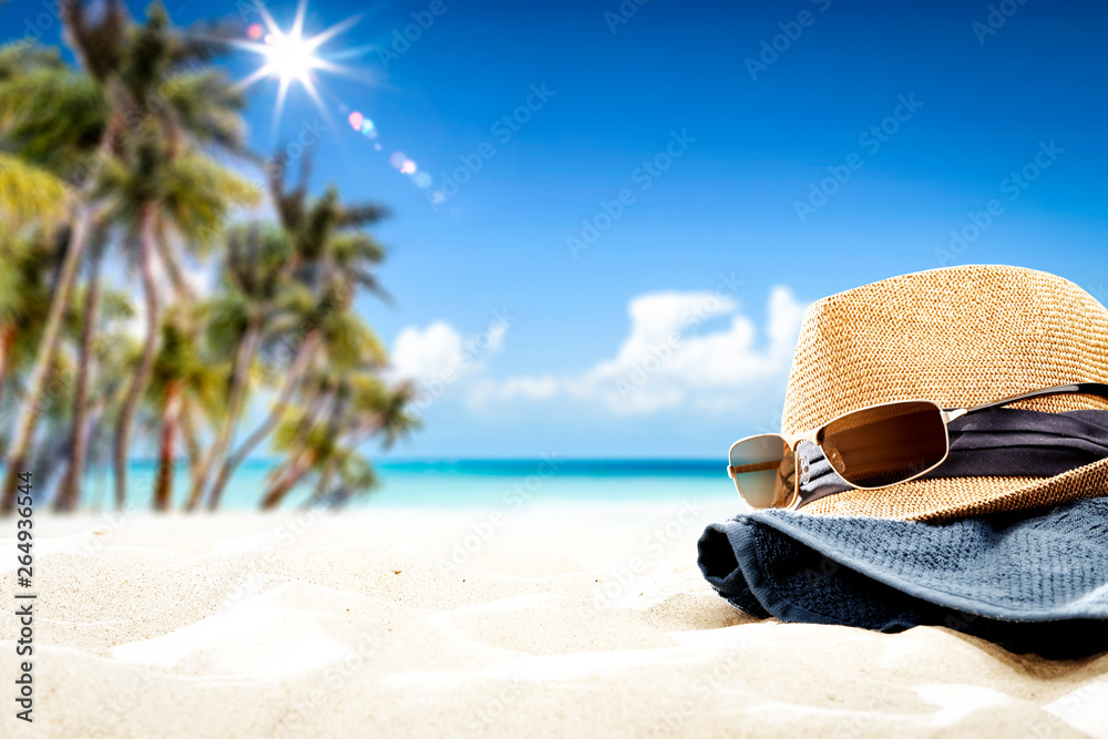 Summer sunglasses with hat on sand and blue towel. Free space for your decoration. Blurred background of beach with palms and ocean Summer sunglasses with hat on sand and blue towel. Free space for your decoration. Blurred background of beach with palms and ocean