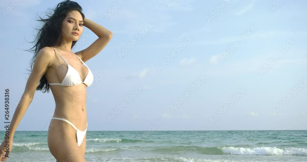 Sexy asian woman in white bikini looks into camera and walks along ocean shore. Steadicam slow motion shot. Summer, Travel and Beauty concept.