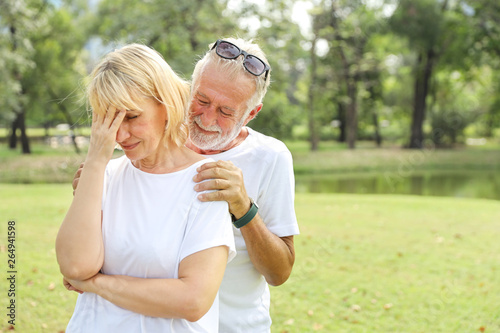 elderly husband is comforting wife and put his hands to her shoulder with love in the park with green tree during summer time
