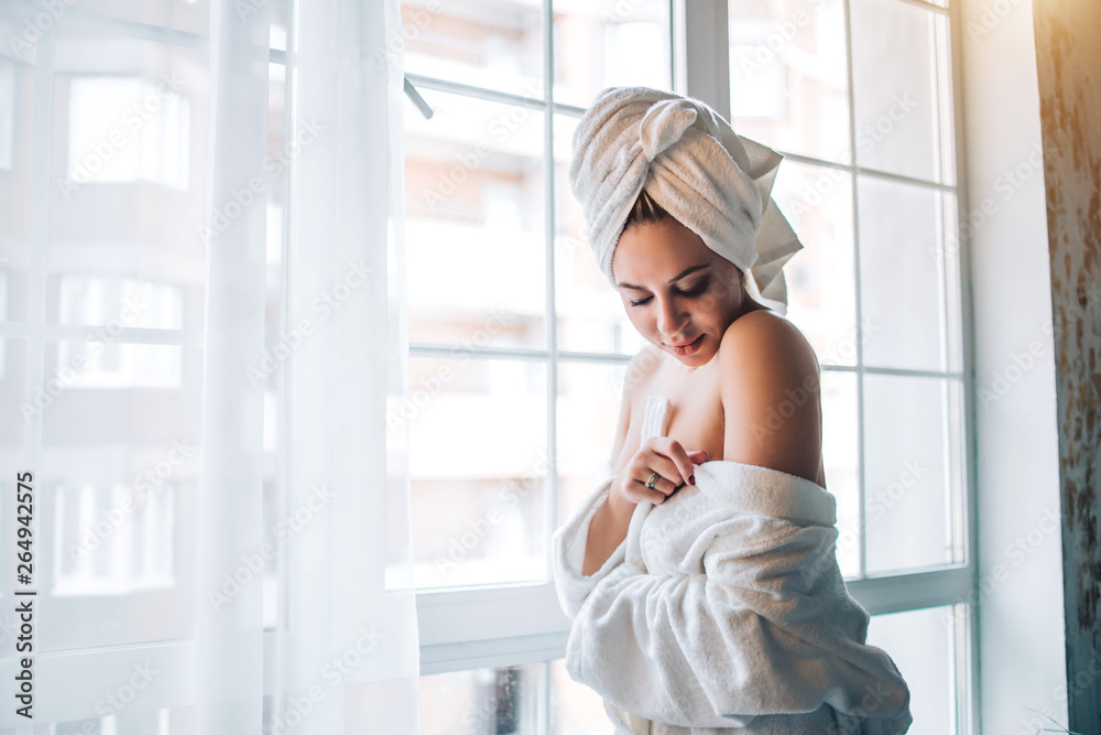 Sensual lovely caucasian shy woman enjoying soft skin, standing in tempting pose near window in hotel room wearing white bathrobe. Purity, tenderness and spa body care concept.