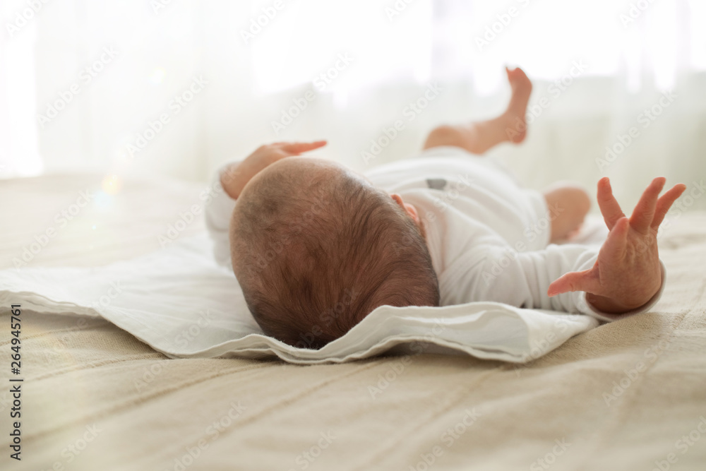 The baby is lying on the bed. A newborn baby looks out the window and moves his arms and legs. Focus the camera on the baby's head.