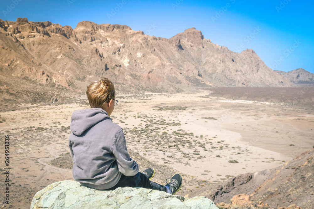 Thoughtful young blonde boy sitting in front of corona forestal of Teide volcano in Tenerife. Cute traveler enjoying great volcanic views. Contemplation, peace, relax, leisure moment concepts