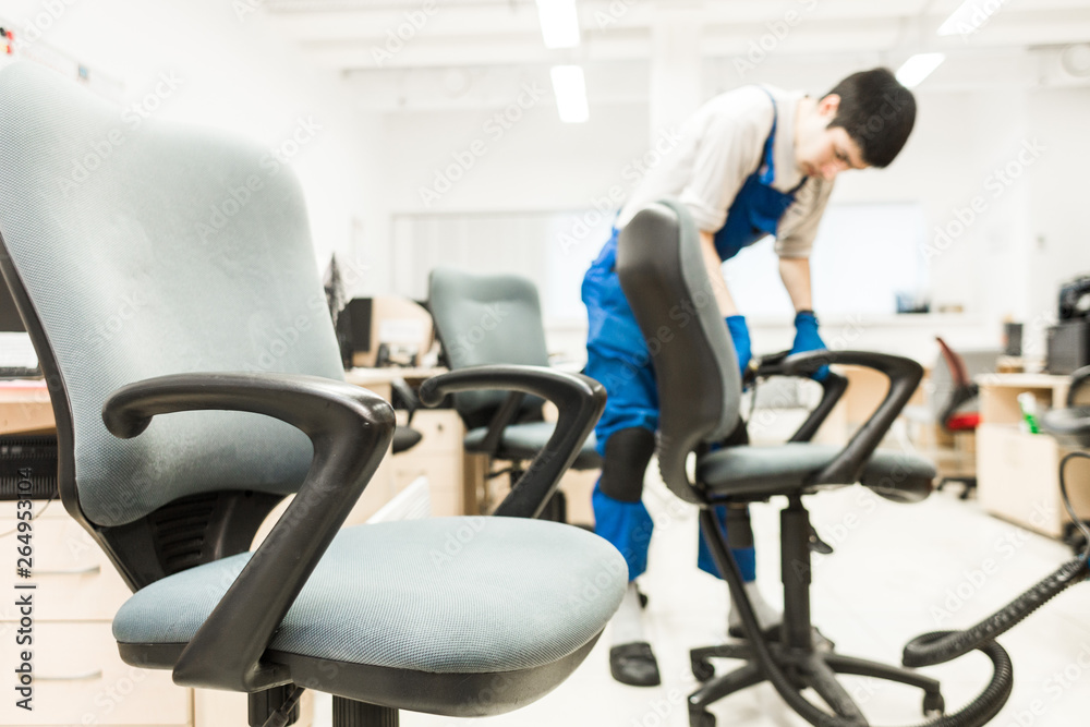 Young man in workwear and rubber gloves cleans the office chair with professional equipment.