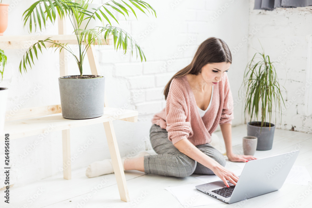 A young happy woman sits on the floor in a bright apartment or office interior and works at a laptop, freelancer girl at work, the concept of distance learning students, paperwork and online learning A young happy woman sits on the floor in a bright apartment or office interior and works at a laptop, freelancer girl at work, the concept of distance learning students, paperwork and online learning