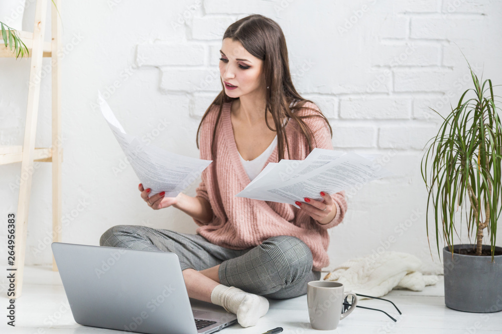 Obraz premium Happy woman examines the paper sitting near the laptop, the girl freelancer works with documents. Student female gets education remotely online