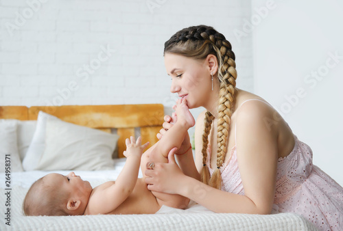 A young mother spends time with her baby.  She plays with him and kisses his little feet. The baby is happy and smiling lying on his back. The baby pulls his hands to mom.