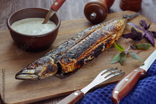 Tasty grilled mackerel on a wooden board next to a white sauce in a ceramic cup, basil sprigs, fork and kinfe and pepper and salt shaker, closeup photo