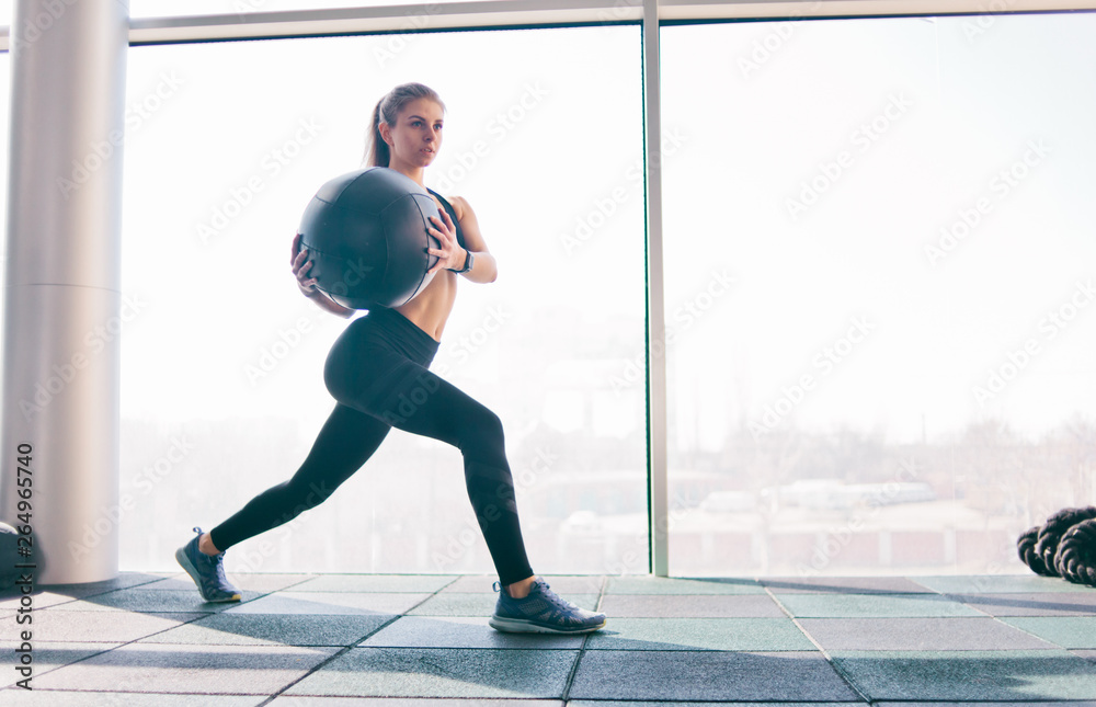 Fototapeta premium Functional training. Fitness young woman training with medicine ball in her hands against the background of a large panoramic window in the gym.
