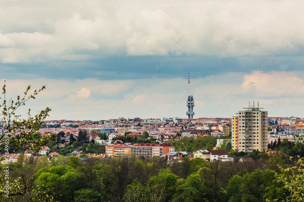 Panoramic view of cityscape of spring Prague, Czech Republic, and its landmark Zizkov television tower. Grey stormy clouds sky, colorful houses and buildings, green vegetation in foreground
