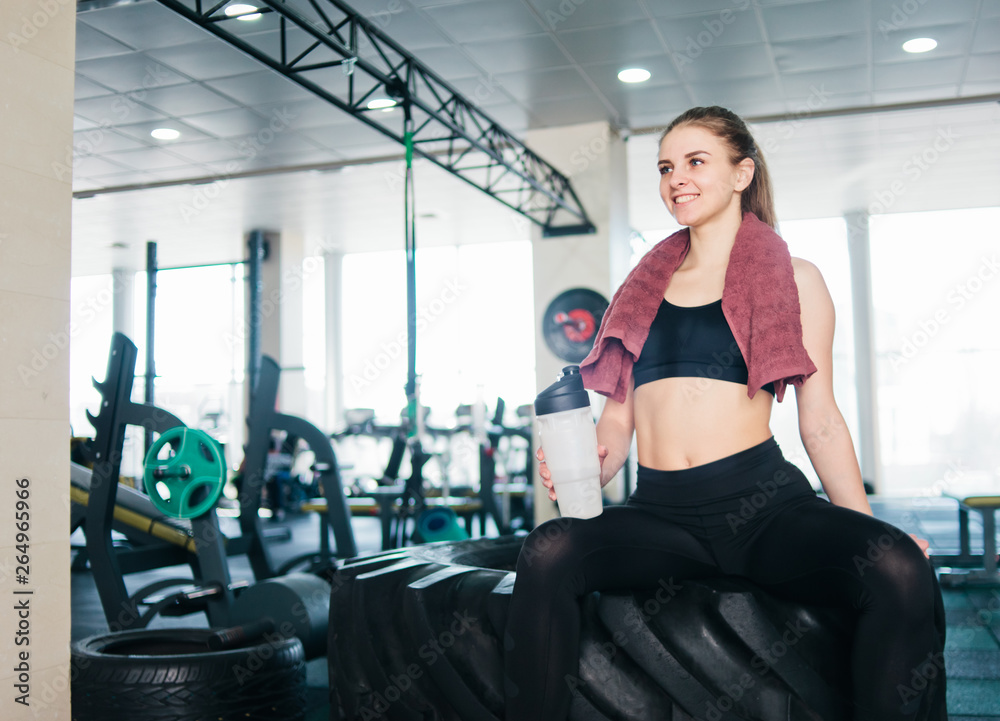 Fototapeta premium Young woman in sportswear with towel around her neck holding bottle of water and sitting resting on the big wheel for functional training in the gym