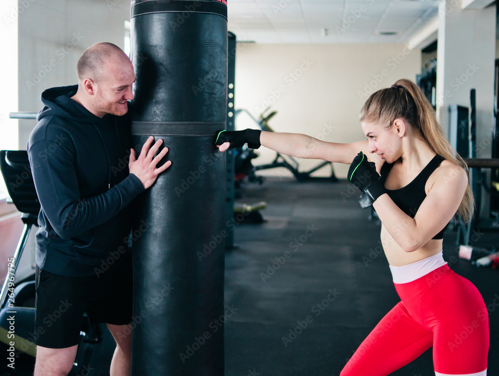 Male instructor trains young woman to do hand punches  at punching bag in the gym.