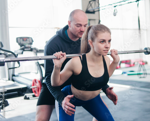 Young sportive woman crouches with a barbell on her shoulders with the help of a male instructor in the gym