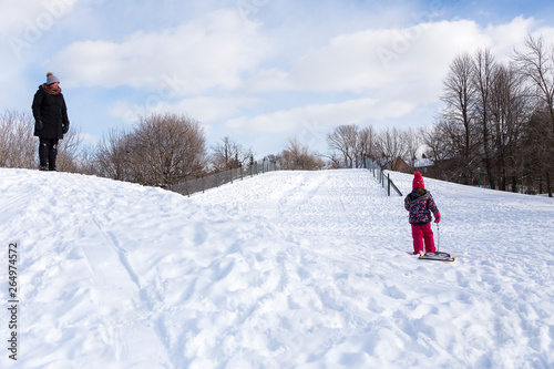 Mother watching little girl with sledge climbing snow-covered hill during a bright sunny winter day, Beaubien Park, Montreal, Quebec, Canada