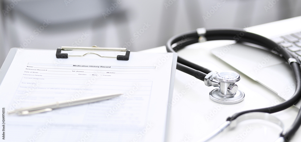 Stethoscope, clipboard with medical form lying on hospital reception desk with laptop computer and busy doctor and patient communicating at the background. Medical tools at doctor working table Stethoscope, clipboard with medical form lying on hospital reception desk with laptop computer and busy doctor and patient communicating at the background. Medical tools at doctor working table