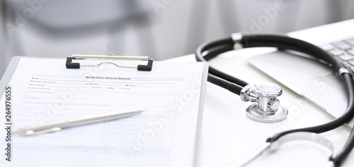 Stethoscope, clipboard with medical form lying on hospital reception desk with laptop computer and busy doctor and patient communicating at the background. Medical tools at doctor working table