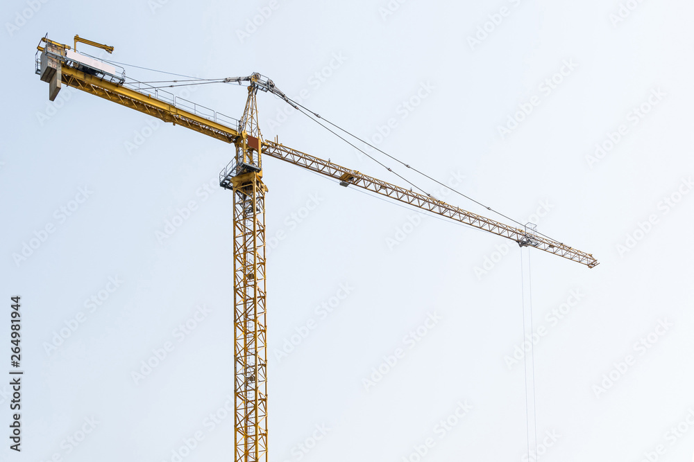 high-rise construction crane with a long arrow of yellow color against the blue sky over a new multi-storey building of concrete and brick under construction
