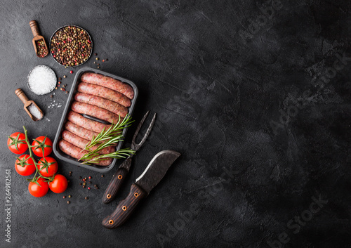 Raw beef and pork sausage in plastic tray with vintage knife and fork on black background.Salt and pepper with tomatoes and rosemary.Space for text