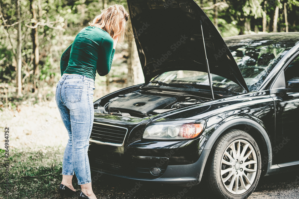 Young woman standing next to a broken car on countryside. Woman's car broken. Woman trying to fix a broken car. Despaired woman trying to fix broken car.