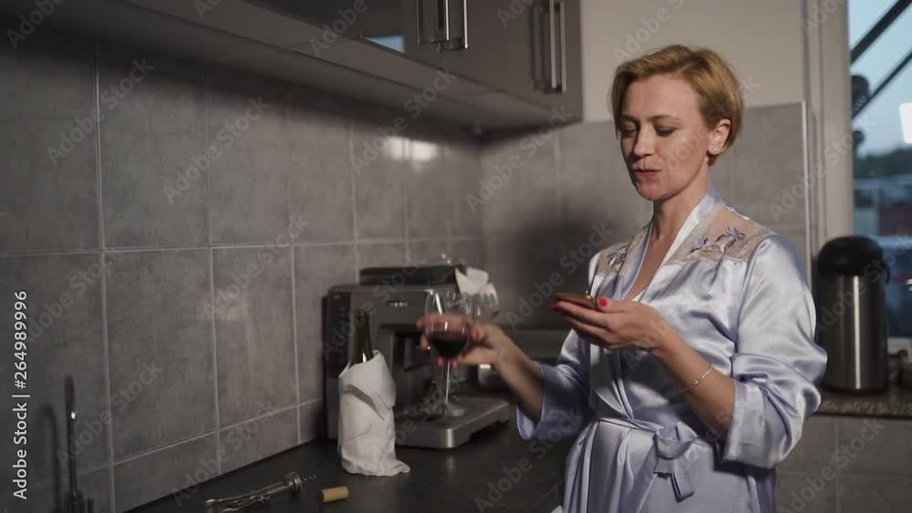 While using her phone Single blonde woman drinking red wine from a glass in a kitchen wearing blue morning gown - Laughing and smiling While using her phone Single blonde woman drinking red wine from a glass in a kitchen wearing blue morning gown - Laughing and smiling