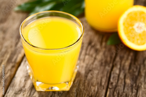 Orangensaft in Glas auf rustikalem Holz mit Orangen im Hintergrund (Selektiver Fokus, Fokus auf die Vorderseite des Glasrandes)