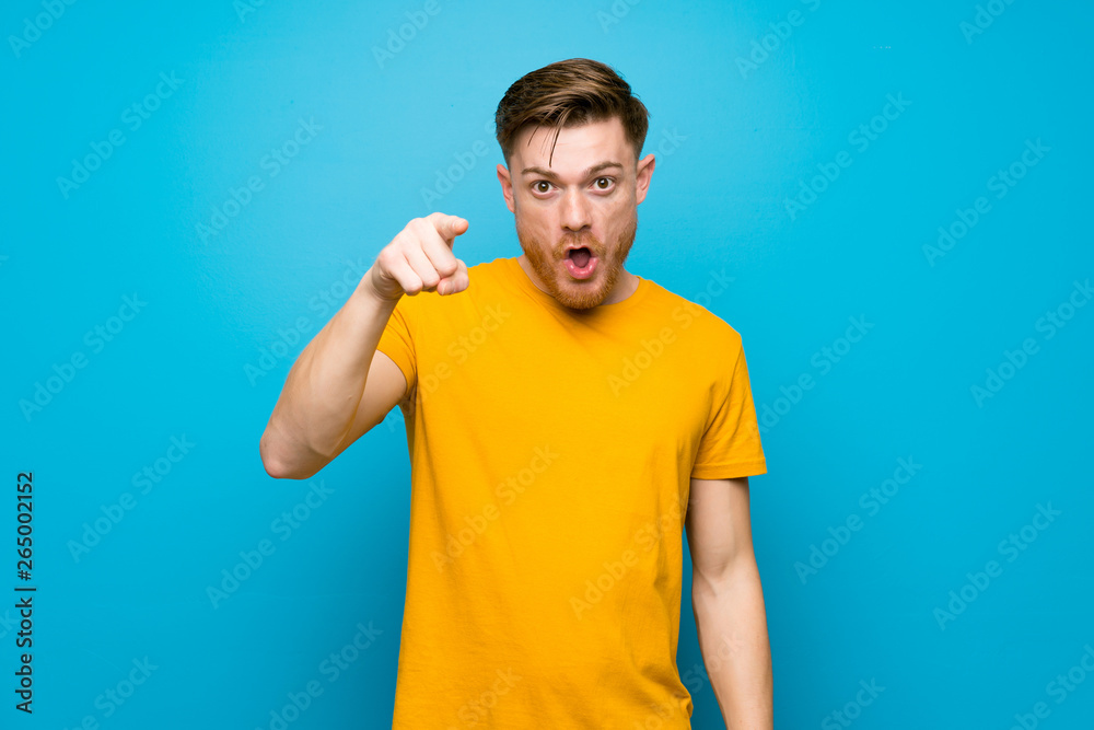 Redhead man over blue wall surprised and pointing front Redhead man over blue wall surprised and pointing front