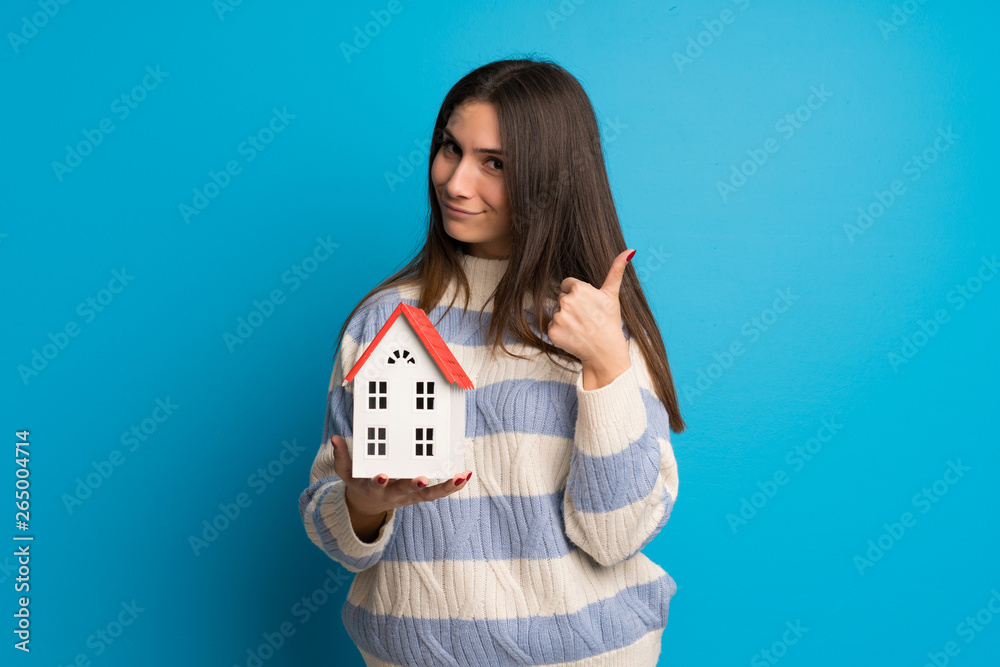 Young woman over blue wall holding a little house Young woman over blue wall holding a little house