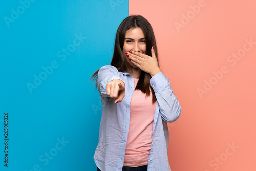 Young woman over pink and blue wall pointing with finger at someone and laughing