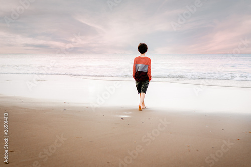 Canvas Print Young boy in striped shirt walking towards the water on the shore gazing at suns