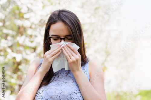 Sick young girl against blooming trees in spring. Outdoor shot of irritated female with seasonal allergy, uses white tissue, poses in light blouse with napkin, has rhinitis and sneezing