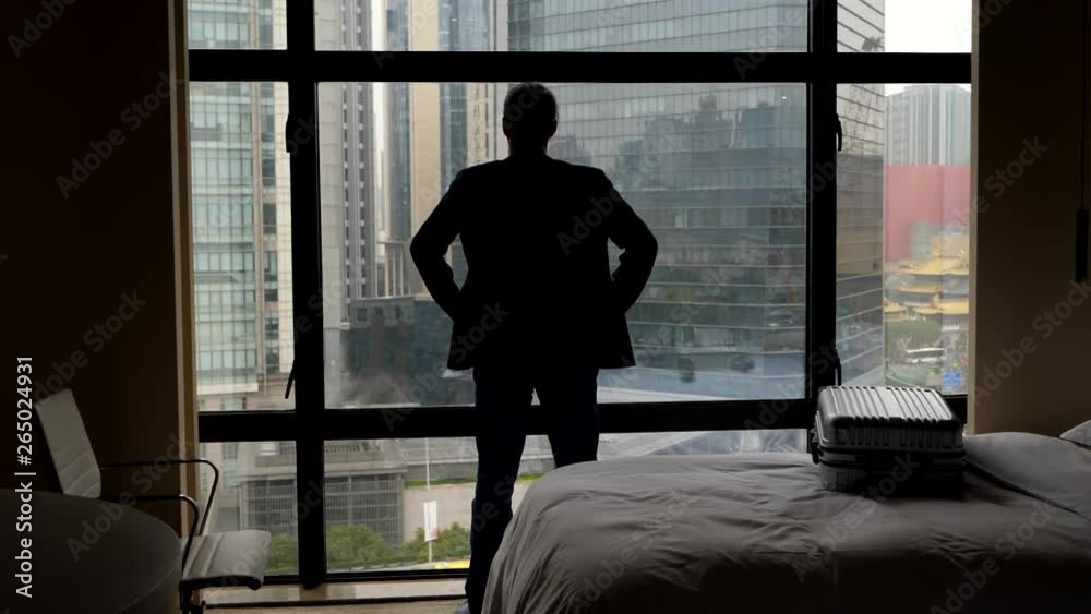 Business man stay alone in bedroom against window, looking out to city buildings. Small closed bag lie on bed. Traveller take pause, thinking or contemplate, standing still with hands on hips Business man stay alone in bedroom against window, looking out to city buildings. Small closed bag lie on bed. Traveller take pause, thinking or contemplate, standing still with hands on hips
