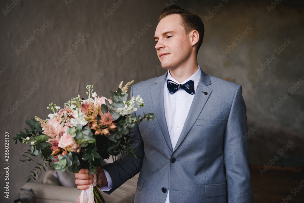 The groom is waiting for the bride in a gray suit. Groom holding bouquet and waiting for bride. Wedding day. The groom stands near a large window. The groom is waiting for the bride in a gray suit. Groom holding bouquet and waiting for bride. Wedding day. The groom stands near a large window.