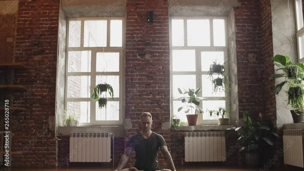 Male practicing yoga in lotus pose indoors. Young man doing yoga in studio with wooden floor and big windows. Freedom, health and yoga concept. Camera slide down with copy space Male practicing yoga in lotus pose indoors. Young man doing yoga in studio with wooden floor and big windows. Freedom, health and yoga concept. Camera slide down with copy space