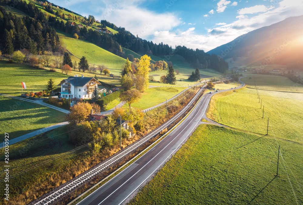 Obraz premium Aerial view of the road in mountain valley at sunset in summer in Dolomites, Italy. Top view of cars on asphalt roadway, house, railroad, hills with green meadows, blue sky, trees, buildings. Highway