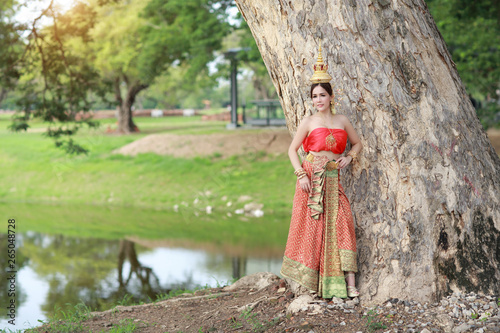 young asian fashion girl wearing Thai traditional costume with beauty face standing beside big green trees in nature outdoors (using for background)