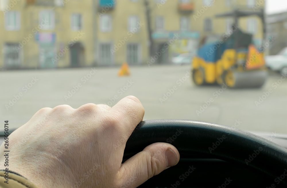 Close-up, the driver's hand on the steering wheel of the car on the background of construction road machinery, road paver and road repair in cloudy weather